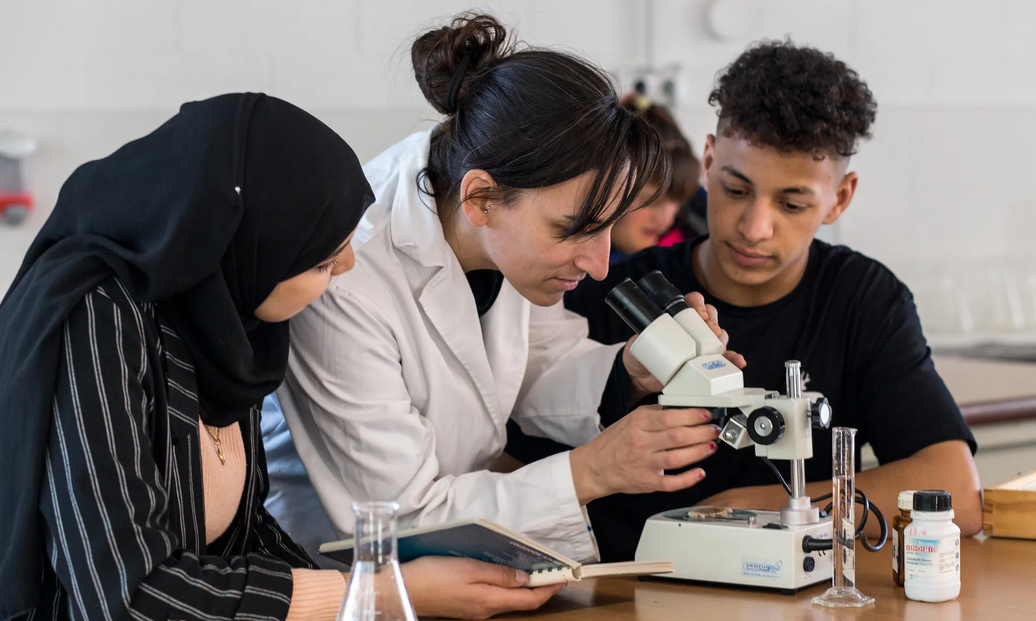 Una professora amb dues alumnes que estan explorant a través d'un microscopi.